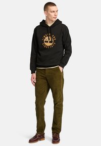 Felpa nera con logo grafico arancione, pantaloni di corduroy verde oliva e scarpe marroni con lacci e suola spessa. Design semplice, vestibilità casual.