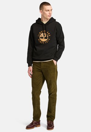 Zwarte hoodie met oranje grafisch logo, olijfgroene corduroy broek en bruine veterschoenen met een dikke zool. Eenvoudig ontwerp, casual pasvorm.