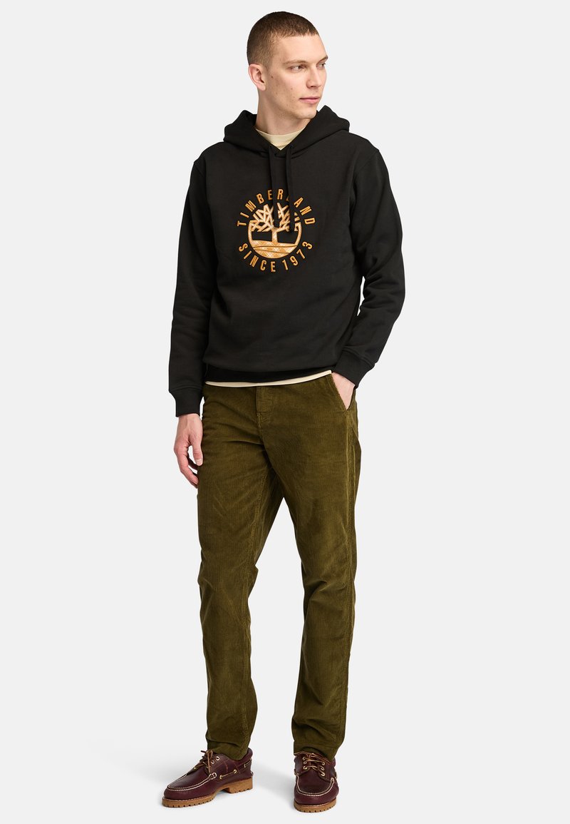 Felpa nera con logo grafico arancione, pantaloni di corduroy verde oliva e scarpe marroni con lacci e suola spessa. Design semplice, vestibilità casual.