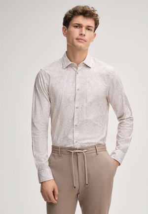 Chemise blanche avec un léger motif floral, manches longues, col à boutons, associée à un pantalon beige avec une taille élastique.