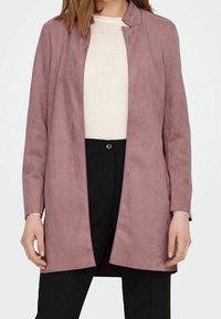 Lange, hellrosa Jacke aus Kunstwildleder mit Stehkragen, offenem Vorderteil und langen Ärmeln. Getragen über einem weißen Oberteil und schwarzen Hosen.