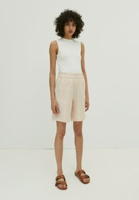 Weißes ärmelloses Oberteil, kombiniert mit hellrosa gestreiften Shorts mit elastischem Bund. Das Model trägt braune Sandalen. Schlichte Hintergrund.