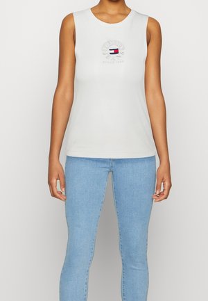 Personne portant un débardeur blanc sans manches avec un logo coloré Tommy Jeans et un jean skinny bleu clair sur un fond uni.