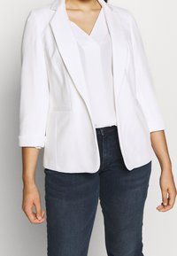 Bílá texturovaná blazerka s klopovým límcem, ohrnutými rukávy a dvěma předními kapsami, nošená přes bílé v-neck tričko a tmavé džíny.