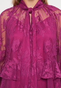 Fuksia blondebluse med blomster mønster, knytehals og volang peplumkant, laget av gjennomsiktig stoff for en delikat tekstur.