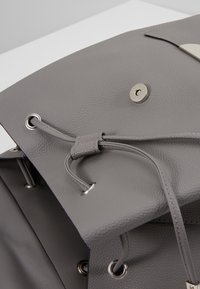 Sac en cuir gris avec fermeture par cordon, œillets métalliques et bouton-pression sur une surface blanche.