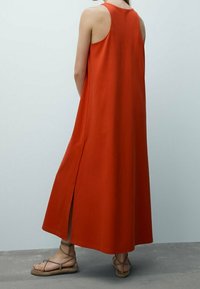 Robe longue sans manches orange avec une coupe décontractée, dotée de fentes latérales. Portée avec des sandales plates à lanières, sur un fond neutre.