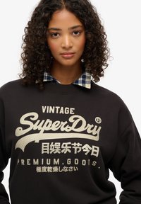 Zwarte sweatshirt met "SuperDry"-logo in goud, voorzien van een gelaagde geruite overhemdkraag in blauw en beige. Het stof lijkt zacht en gezellig.