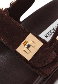 Bruine suède sandaal met een rechthoekige gouden gespsluiting. Heeft een zachte textuur en gestikte accenten rond de band.