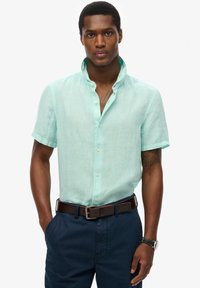 Uomo con indosso una camicia azzurra chiaro a maniche corte con le maniche arrotolate, pantaloni blu scuro, cintura marrone e orologio da polso nero, in piedi con le mani in tasca.