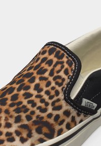 Chaussure à enfiler avec tige imprimé léopard, bordure noire, couture blanche et logo Vans sur le côté intérieur près du talon.