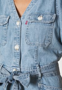 Vaaleansininen farkkahaalari, jossa on V-pääntie, kaksi etutaskua, valkoiset napit ja sidottava vyö, joka tuo lisää muotoa.