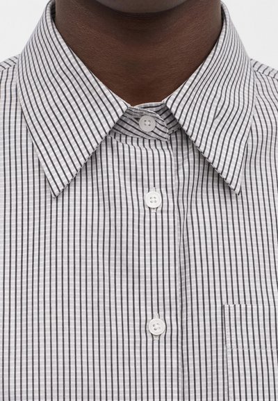 Κοντινό πλάνο ενός ατόμου που φοράει λευκό πουκάμισο με λεπτές μαύρες κάθετες ρίγες και λευκά κουμπιά, δείχνοντας το γιακά και την πάνω περιοχή του στήθους.