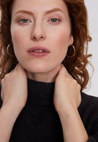 Zbliżenie na osobę z kręconymi, średniej długości czerwonymi włosami. Ubrana w czarny sweter z golfem i złote kolczyki w kształcie kół, z lekkim makijażem.