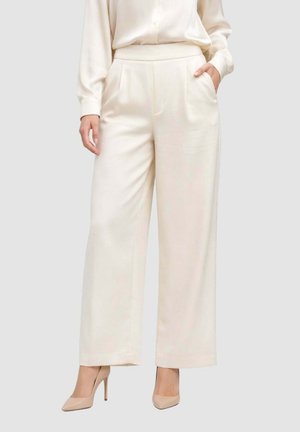 Donna che indossa pantaloni crema a vita alta e gamba larga con tasche laterali, abbinati a una camicetta crema e scarpe con tacchi alti color nude.