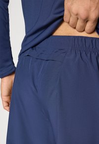Navy athletic pants with a textured fabric and elastic waistband. Side pockets and a slight sheen are visible near the waistband area.