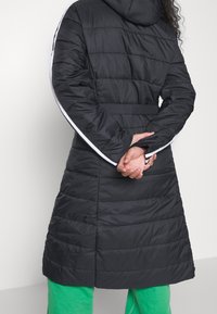 Abrigo acolchado negro con capucha, que cuenta con costuras horizontales, acentos laterales blancos y una cintura ceñida con cinturón. Fabricado con un material cálido y ligero.