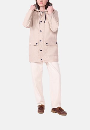 Person trägt beige Kapuzenjacke mit vorderen Knöpfen und Taschen, weiße Hose und braune Schnürschuhe vor einfarbigem Hintergrund.