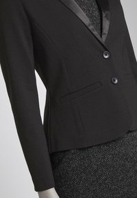 Blazer nero con rever in satin, due bottoni e tasche laterali, indossato sopra una gonna nera tessurizzata, che mette in risalto una silhouette aderente.