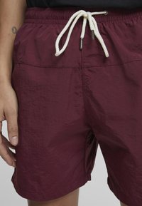 Burgunderfarbene Badeshorts aus leichtem Stoff, mit strukturierter Oberfläche, elastischem Bund und weißem Zugband mit metallic-farbenen Enden.