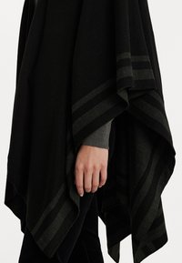 Melnā pončo ar tumši zaļām svītrām, izgatavots no teksturēta auduma, eleganti nolikts uz rokas, izceļot slānveida dizainu.