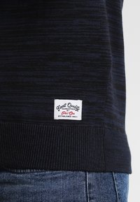 Ciemnoniebieski sweter dzianinowy z subtelnym wzorem poziomych pasków, prążkowanym dołem i białą metką z napisem "Najwyższa Jakość" oraz "Pie's One".