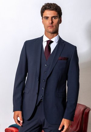 Young man wearing a tailored dark blue three-piece suit with a white shirt and burgundy tie and pocket square, standing against a plain background.
