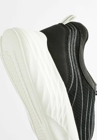 Black athletic shoe with textured fabric upper, white rubber sole, and side accents. Features a pull tab on the heel for easy wear.