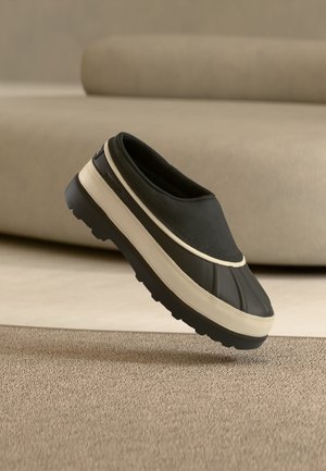 Μαύρο και μπεζ αδιάβροχο παπούτσι χωρίς κορδόνια με χοντρή σόλα που ακουμπάει σε μπεζ χαλί μπροστά από μπεζ καναπέ.