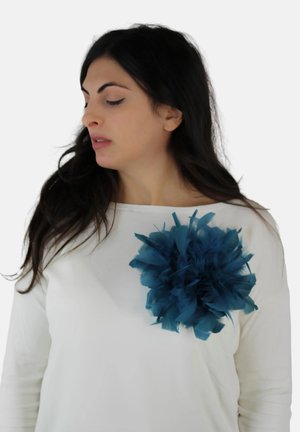 Vrouw met lang donker haar, gekleed in een wit shirt met een grote blauwe stoffen bloem op de linkerborst, kijkt naar links.