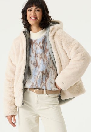 Lachende vrouw die een lichtbeige donzige jas draagt, een trui met dierenprint in blauw en bruin, beige broek en een lichtbruine riem met gouden gesp.