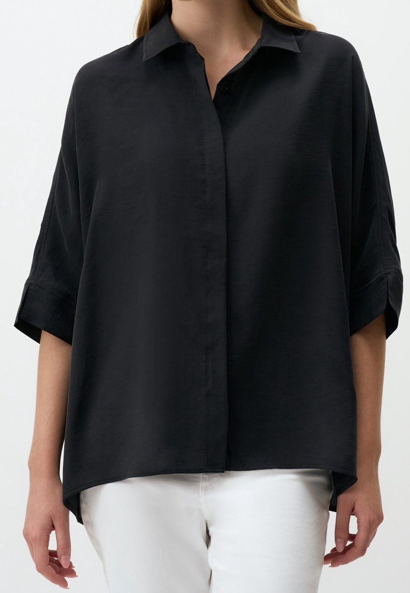 Femme portant une chemise noire ample à boutons avec manches retroussées et un pantalon blanc sur un fond uni.
