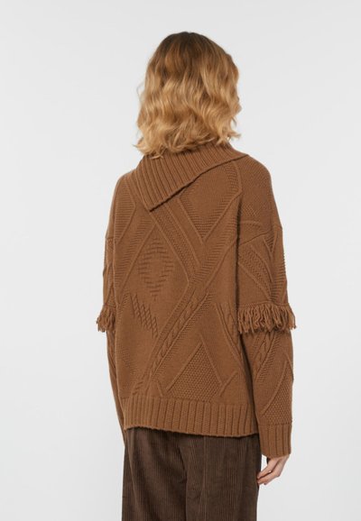 Una donna con capelli mossi di lunghezza spalla indossa un maglione di lana marrone con motivi geometrici e frange sulle maniche, abbinato a pantaloni in velluto a coste marroni.