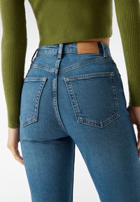 Höga blå jeans med bakfickor. Denimtyget har en slät struktur. En brun läderetikett är synlig vid midjan.