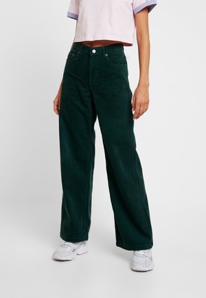 Osoba ubrana w ciemnozielone szerokie spodnie sztruksowe, krótki różowy top z krótkim rękawem i białe trampki, stojąca na jednolitym tle.