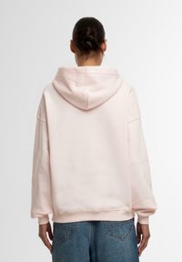 Lys rosa hettegenser med løs passform, ribbede mansjetter og bunn. Mykt stoff, ingen synlige mønstre eller maskinvare detaljer. Brukt med blå jeans.