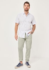 Weißes kurzärmeliges Hemd mit Knöpfen und vertikalen Streifen, kombiniert mit hellgrünen Hosen und blauen Turnschuhen. Auffällige Tattoos an einem Arm.