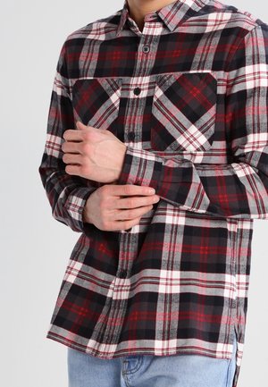 Homme portant une chemise à carreaux noire, rouge, blanche et grise, boutonnée, ajustant le poignet avec un jean bleu clair visible.