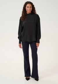 Maglia nera a maniche lunghe con collo alto, realizzata in un tessuto morbido, abbinata a jeans svasati scuri e scarpe nere con punta a mandorla.