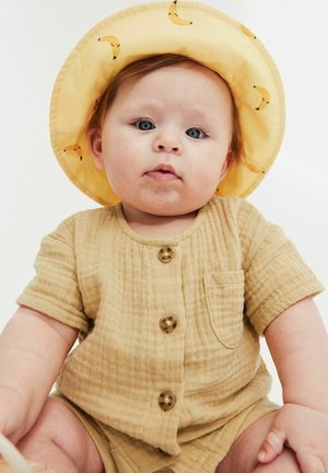 Bébé aux yeux bleus portant une combinaison beige à boutons et un chapeau de soleil jaune avec des imprimés bananes souriantes, assis sur un fond blanc.