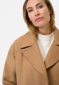 Beige wollen jas met brede revers en een gestructureerde afwerking, gelaagd over een witte gebreide coltrui. Close-up van het bovenlichaam.