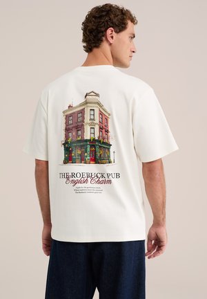 Mann trägt weißes T-Shirt mit bunter Illustration eines dreistöckigen Pubs und dem Text "The Roebuck Pub English Charm" auf dem Rücken.