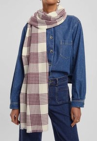 Sciarpa scozzese color crema con accenti marroni, realizzata in morbido tessuto, con orli frangiati, appoggiata su una giacca di jeans.