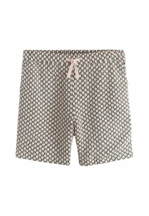 Man die beige en zwart geruite textuur korte broek met trekkoord draagt, staand buiten bij een stenen muur in het zonlicht.