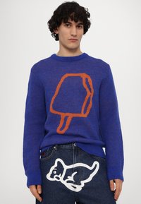 Sweater azul com gráfico de gelado laranja, punhos e gola canelados; combinado com uns jeans escuros que apresentam um gráfico de gato branco na parte de trás.