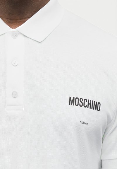 Κοντινό πλάνο ενός ατόμου που φοράει λευκή πόλο μπλούζα με τρία κουμπιά και το λογότυπο "MOSCHINO Milano" τυπωμένο μαύρο στο στήθος.