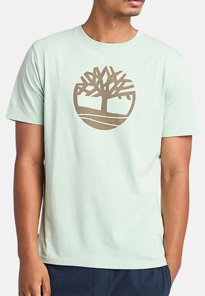 T-shirt verde menta a maniche corte in cotone, con un grande logo di un albero marrone al centro e un elemento di design rotondo sotto di esso.