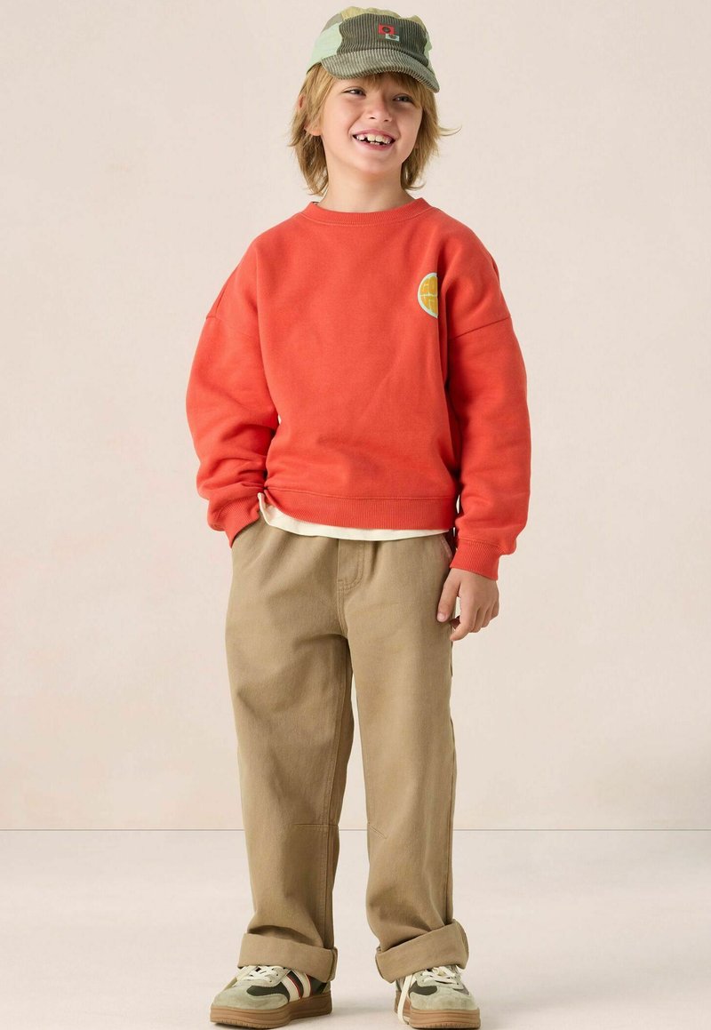 Sweatshirt rouge, pantalon beige, casquette verte et beige avec logo. Les baskets présentent plusieurs couleurs. Texture lisse, coupe décontractée.