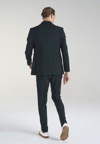 Ciemnozielony, dopasowany garnitur z prostymi spodniami i jednorzędową marynarką. Białe sneakersy stanowią kontrast. Gładki materiał, minimalny design.