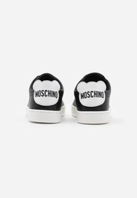 Baskets noires et blanches vues de dos avec des patchs en forme d'ours blancs sur le talon, portant le texte « Moschino » en noir.